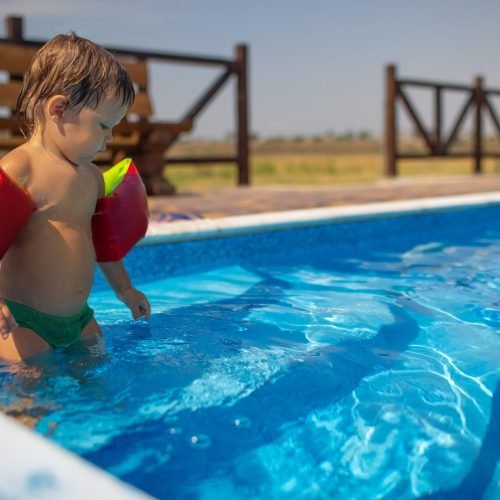 Little boy with bright armbands, playing with beach colored toys near a deep blue pool with clear transparent water against the background of a warm sunny summer sunset